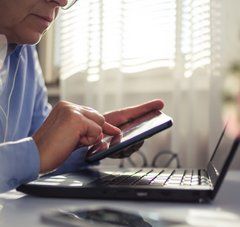 personne assise à un bureau tenant un smartphone dans le main devant un écran d'ordinateur portable